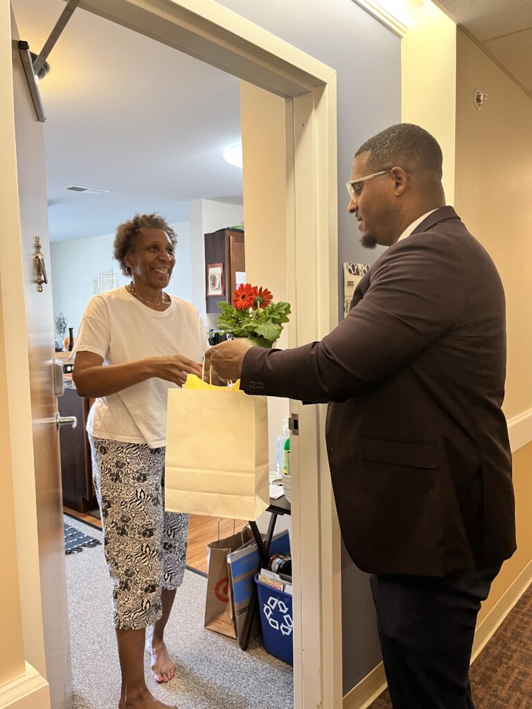 Dr. Junior Dillion hands an older woman a pink potted flower and a white gift bag for "I Remember Mama" at Cobblestone Place apartments.