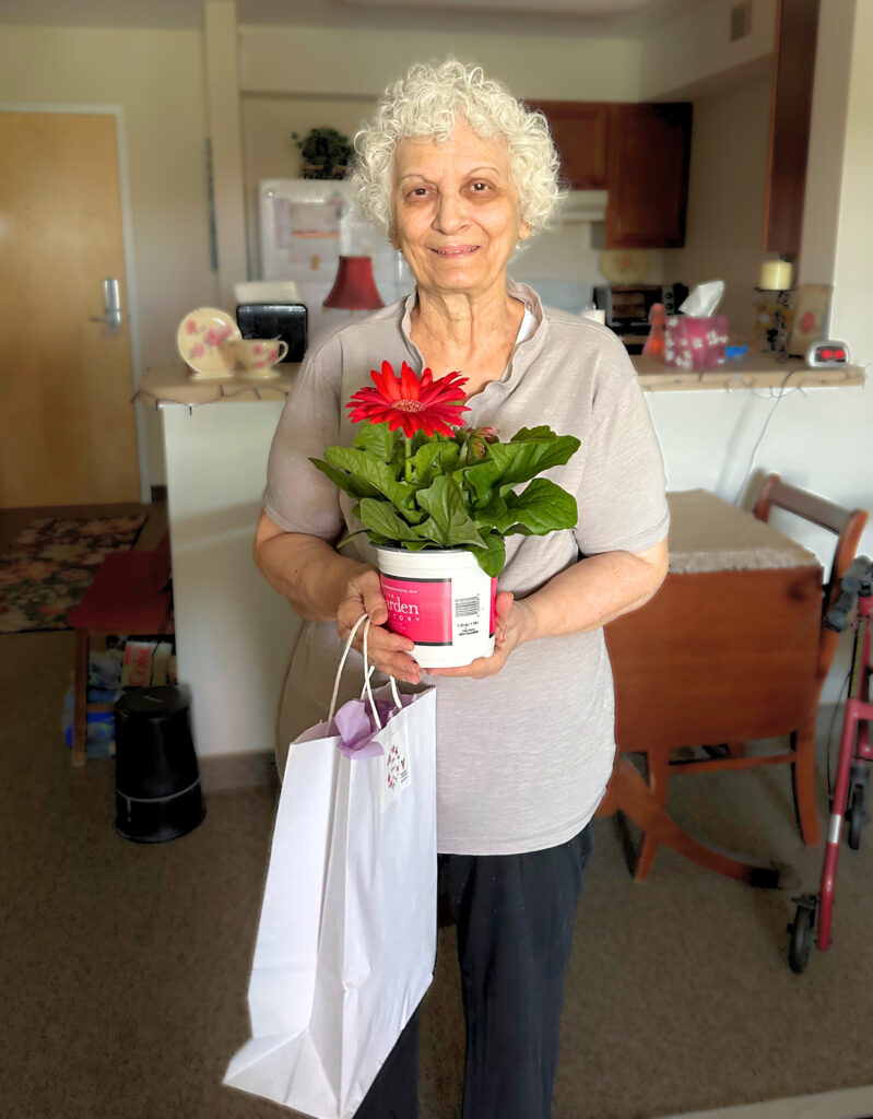 Older woman holds a pink potted flower and a white gift bag for "I Remember Mama" at Cobblestone Place apartments.