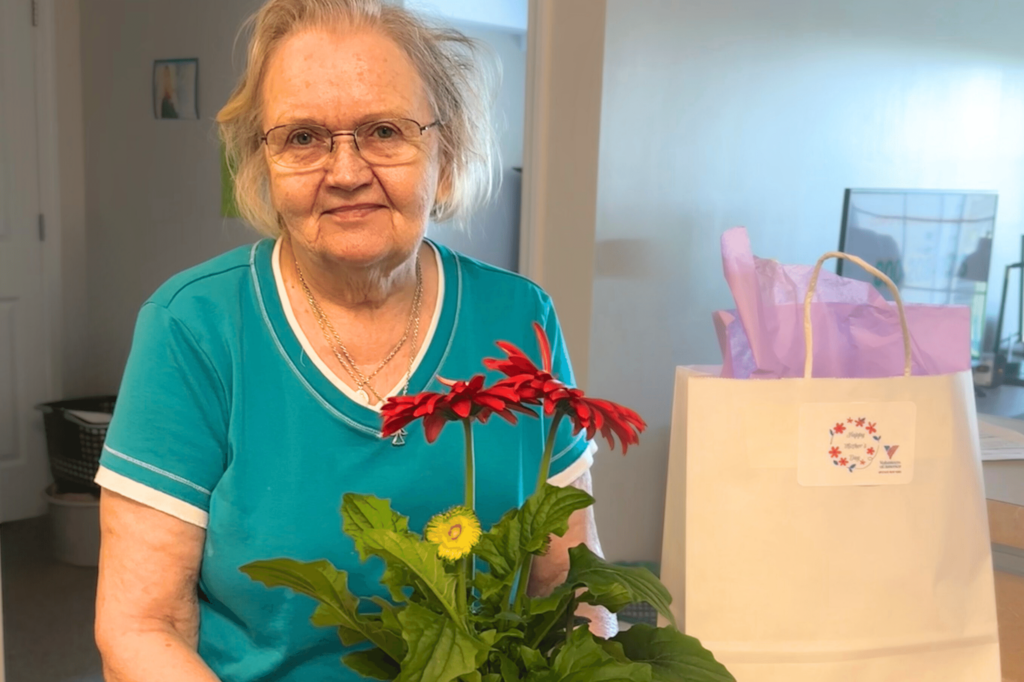 Older woman with white hair and teal shirt stands smiling next to a pot of pink flowers and white gift bag.
