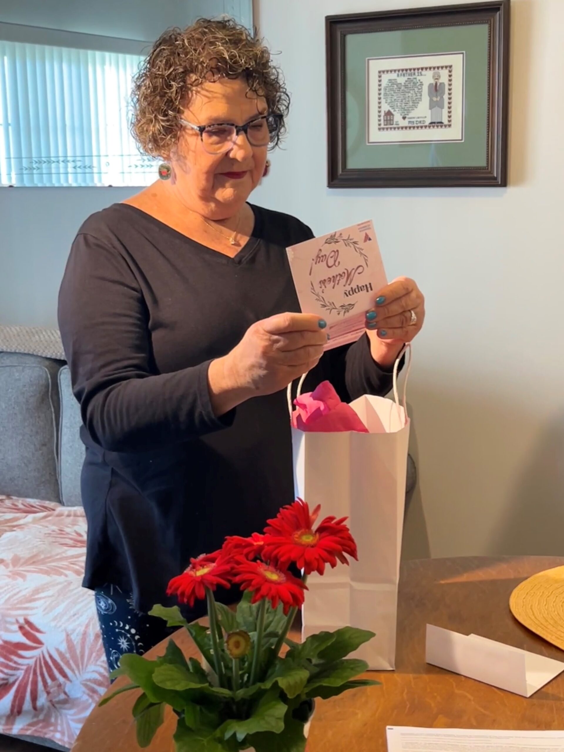 Older woman reads a card with a white gift bag and pink potted flower on the table next to her.