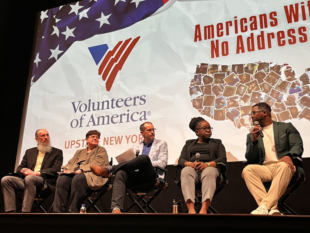 Five people sit on stage in front of a screen with VOA Upstate New York logo as they discuss the "Americans with No Address" documentary