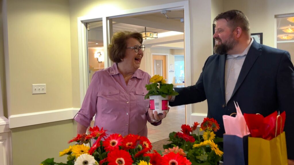 Man hands smiling older woman a potted flower and gift bag at VOA's I Remember Mama tea at Cobblestone Place apartments.