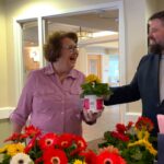 Man hands smiling older woman a potted flower and gift bag at VOA's I Remember Mama tea at Cobblestone Place apartments.